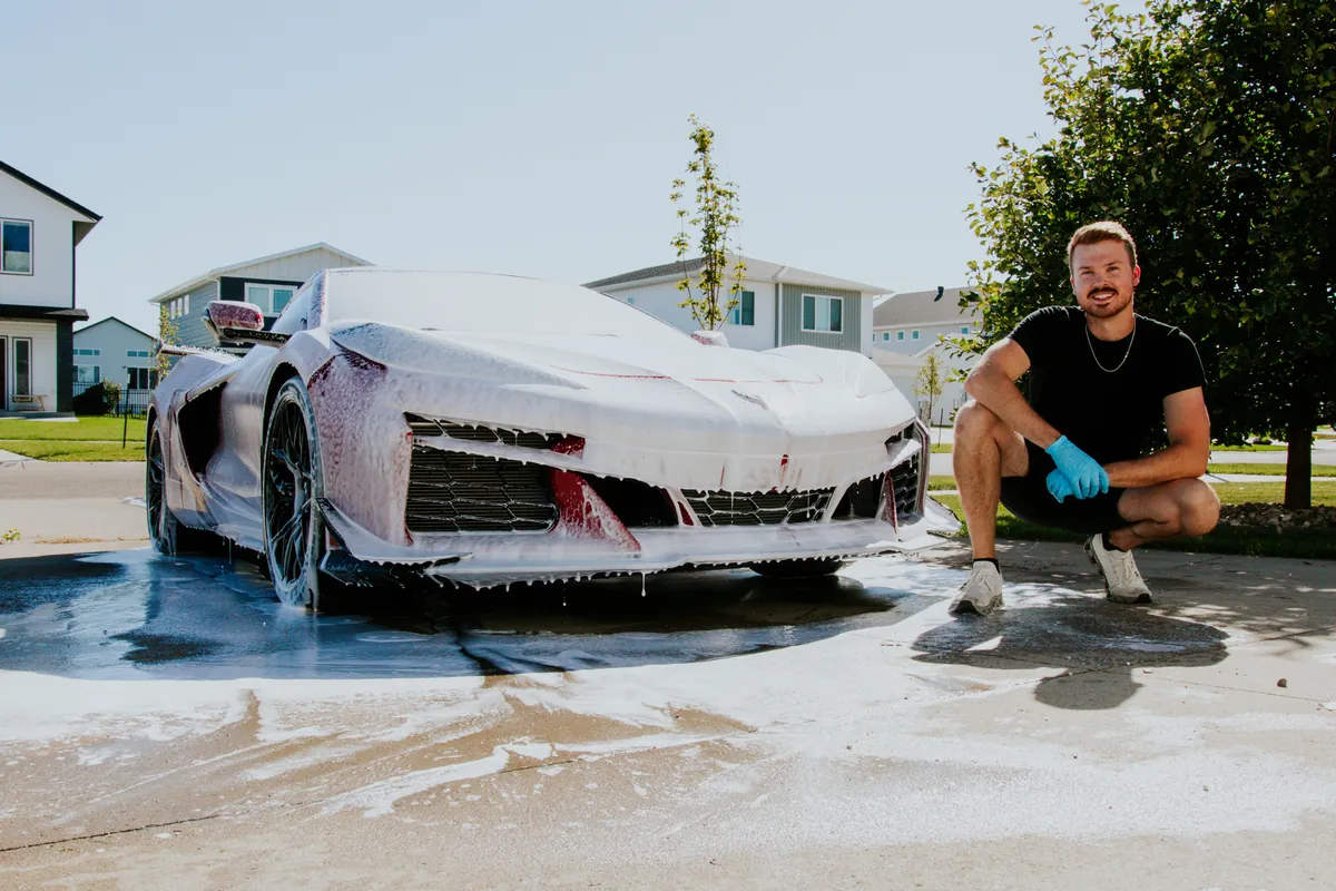 Ryan, owner of WAKE Detail Co., crouching beside a foam-covered Corvette during a mobile detail